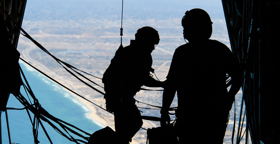 Two military members inside an aircraft in flight with the back ramp open. One member is attached to a cable.