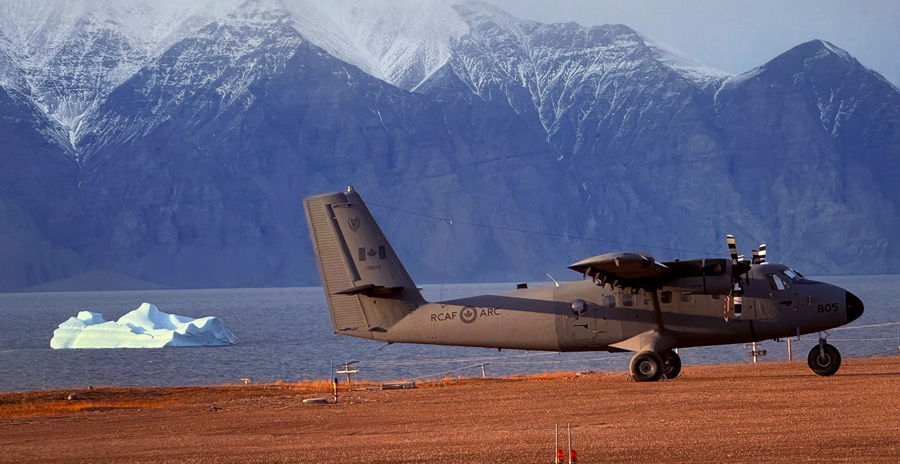 A military aircraft is parked on a gravel runway near a body of water. In the background, there is an iceberg and mountains.