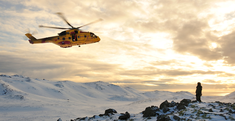 Yellow rescue helicopter flies over snowy mountains as a person stands on a ridge during a training exercise.