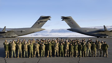Un groupe de membres du personnel en uniforme est debout sur un terrain d’aviation entre deux gros avions de transport militaire. Des collines enneigées sont aperçues en arrière-plan.