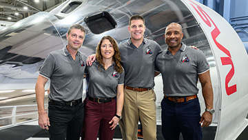 Four people, three men and one woman, stand in front of a NASA Space Launch System crew capsule. They stand close together with their hands on each other’s shoulders.