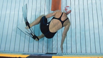 Un nageur portant des palmes, un bonnet de bain et un maillot de bain une pièce foncé est sous l'eau dans une piscine, étendu près du fond.