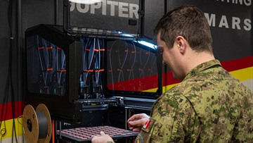 Une personne vêtue d’un uniforme militaire est debout devant une grande imprimante 3D industrielle. Elle tient un plateau rectangulaire rempli d’objets circulaires rouges espacés uniformément. Plusieurs bobines de filament sont fixées sur le côté de la machine.