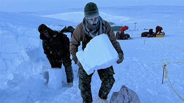 A group of people working outside in the snow and in the foreground two individuals are holding blocks of snow to build a shelter.