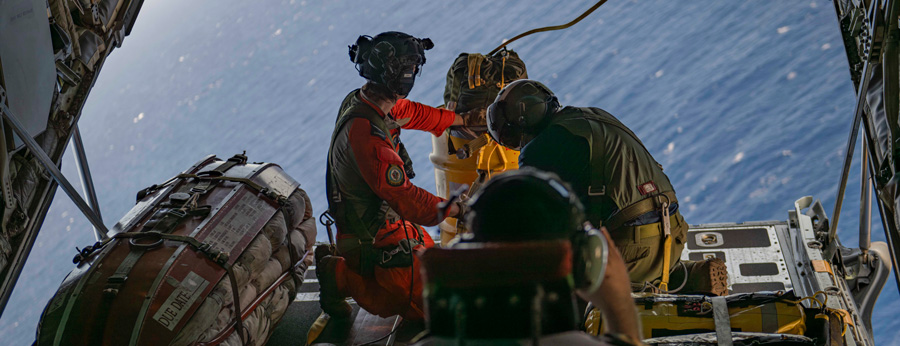 Crew members prepare an airdrop from an open aircraft ramp over the ocean, handling cargo and safety gear.