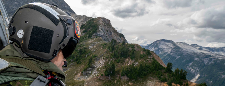A helmeted crew member looks out from a helicopter over rugged, forested mountains under a cloudy sky.