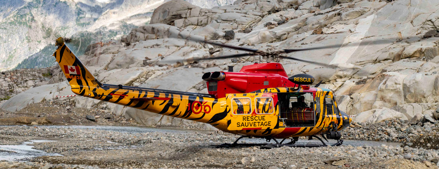 A yellow and red search and rescue helicopter with tiger stripes is parked on rocky terrain with mountains in the background.