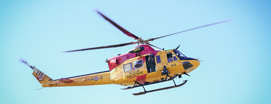 A yellow and red search and rescue helicopter flies against a clear blue sky with its side door open.