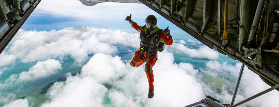 A search and rescue parachutist in orange gear jumps from an aircraft into the sky above clouds and green terrain below.