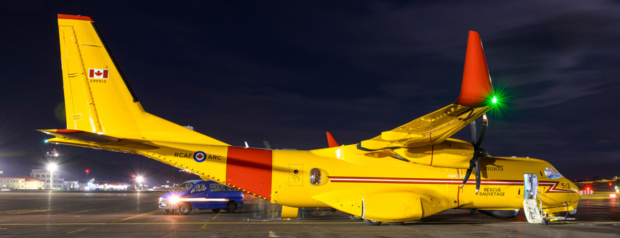 A yellow and red Canadian search and rescue aircraft is parked on a lit tarmac at night with its propellers still.