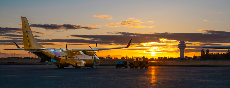 A yellow search and rescue aircraft sits on the tarmac at sunrise with an airport control tower in the background.