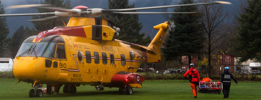 A helicopter is parked on a field and two search and rescue personnel walk towards it carrying a crate filled with supplies.