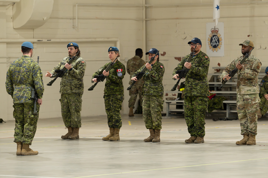 Des membres de l’ARC et un membre de l’United States Air Force se tenant côte à côte tiennent un fusil C7 devant leur poitrine dans un gymnase, tandis qu’un stagiaire debout devant eux leur crie des ordres.