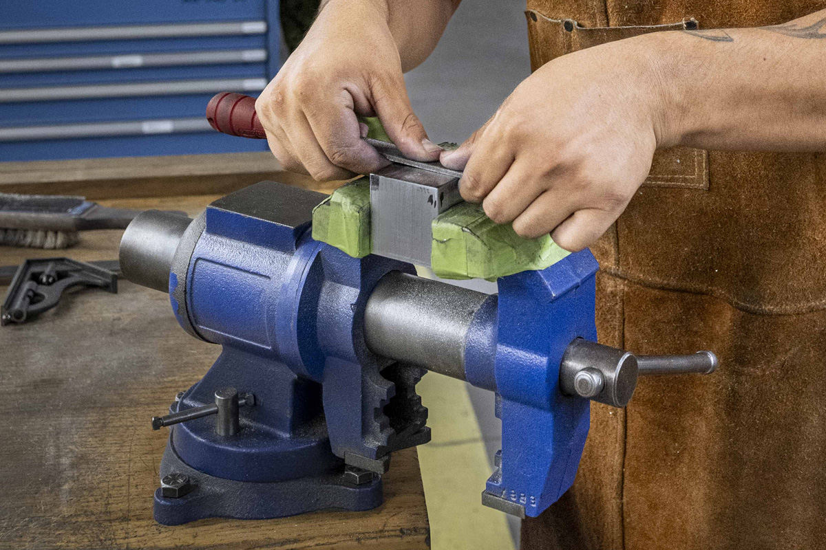 A person in brown apron, wearing safety glassed stands over a vice holding a block of metal while manipulating a file.
