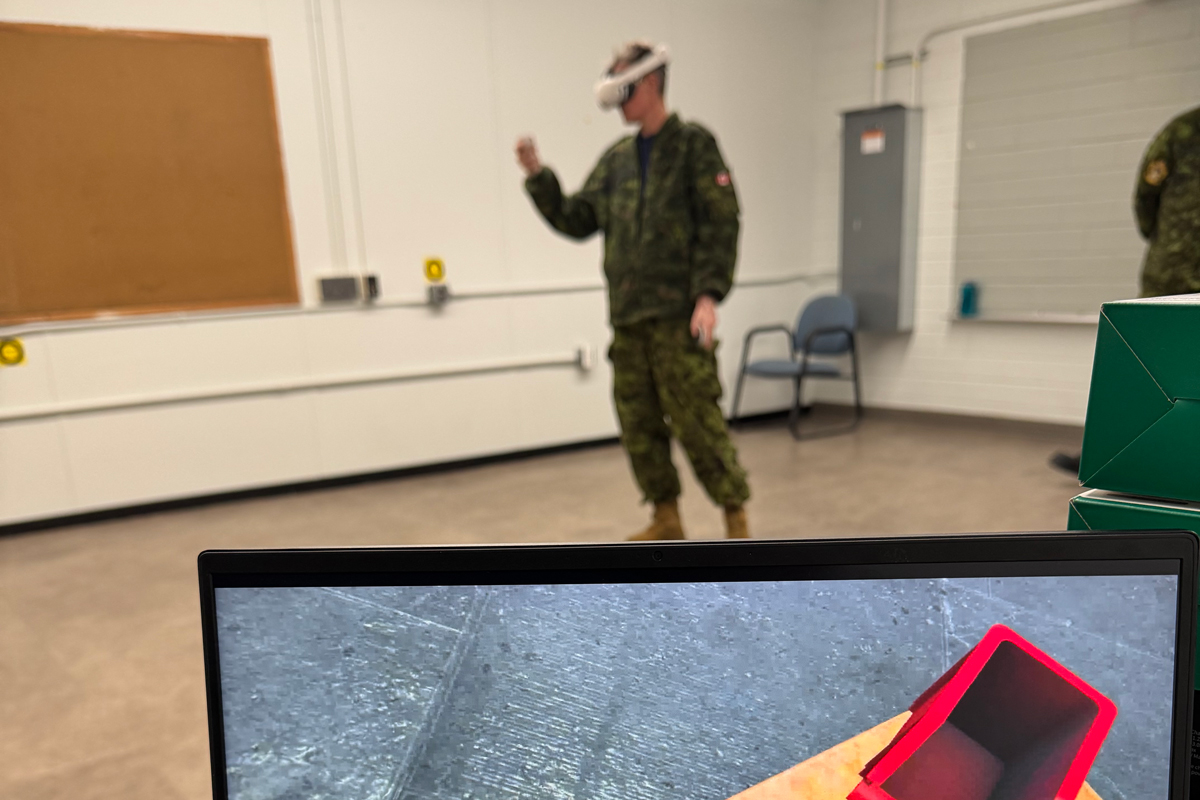 A computer screen is seen on a desk displaying a virtual workbench with a simulated hand holding a u-shaped aircraft component. In the background, a Royal Canadian Air Force member in a camouflage uniform wearing a virtual reality headset stands in a classroom with their hand raised, mimicking what is seen on the screen.