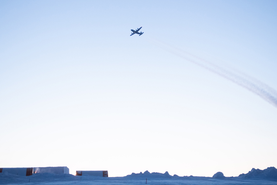 A four-engine aircraft is silhouetted against the sky with condensation trails behind. Below three rectangular buildings with orange stripes are surrounded by snow.