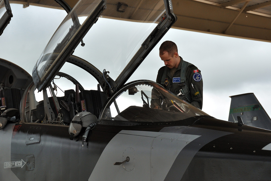 A Royal Canadian Air Force member in a green flight suit looks into the open cockpit of a grey training jet parked next to another jet to the right, under an overhang.