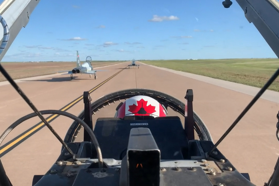 A pilot wearing a helmet painted with the Canada flag is seen from the rear in a cockpit, while two grey jets taxi in front of them along a wide paved surface lined with grass under a blue sky.