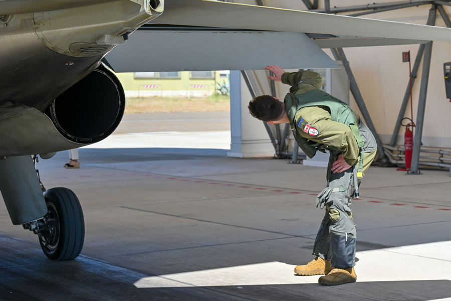A pilot in a flightsuit looks under the wing of a grey aircraft in a hangar.
