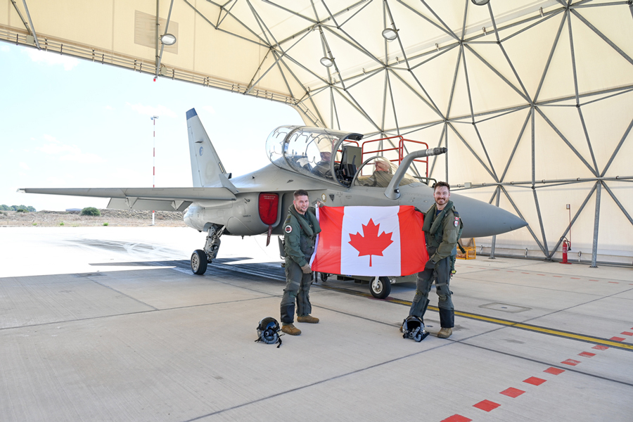 Two pilots in flightsuits hold a Canadian flag in front of a small grey training jet aircraft under a canvass hangar.