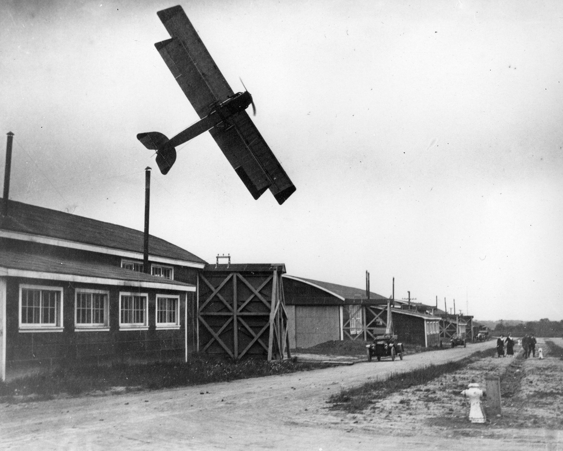 In a black and white photograph, a vintage bi-plane flies extremely low over one-storey wooden buildings.