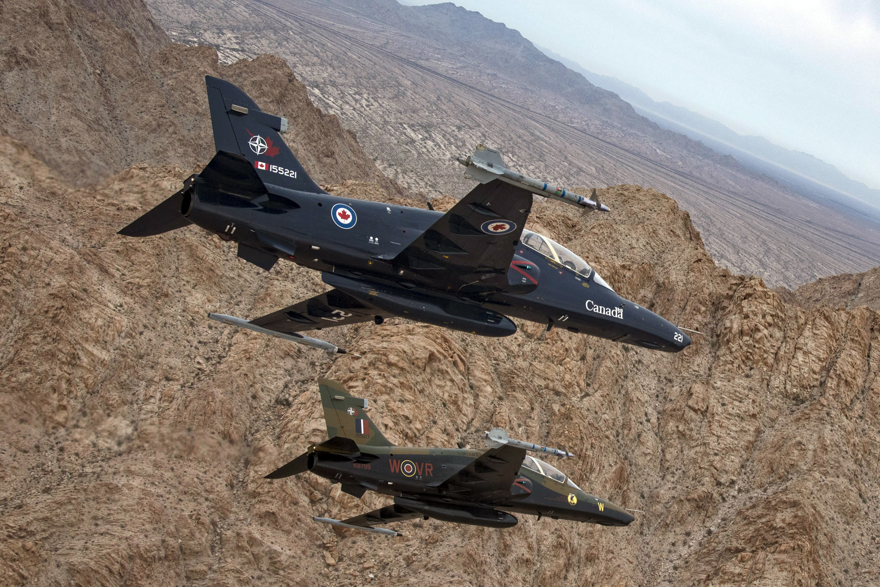 Two aircraft fly side by side over a craggy desert landscape.