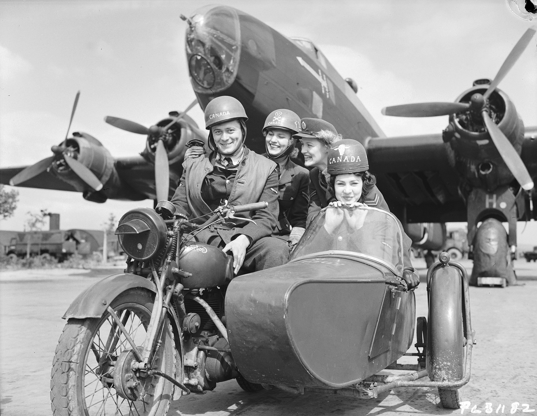 In a vintage black and white photo, four people in military uniforms are seated on a motorcycle or in its sidecar in front of a large aircraft.