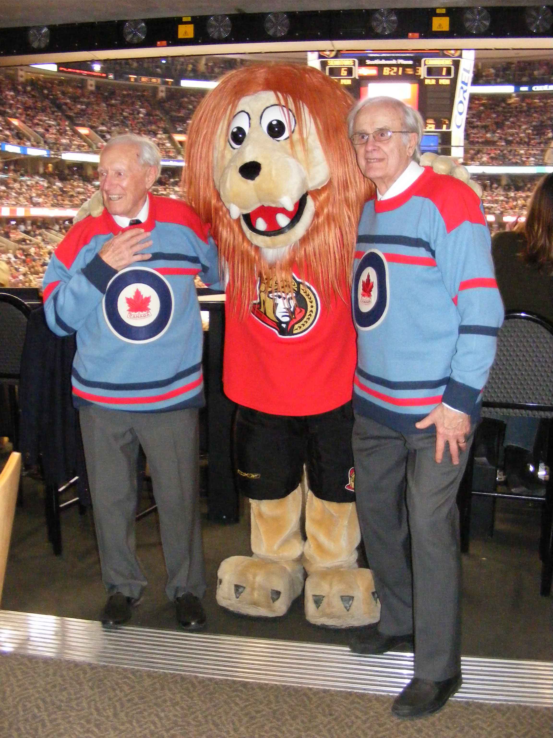 Former RCAF Flyers team mates Ab Renaud (left) and André Laperrière, sporting their replica 1948 Olympics jerseys specially produced by the Royal Canadian Air Force Association, are greeted by Spartacat, the mascot of the Ottawa Senators hockey team. Mr. Renaud and Mr. Laperrière dropped the puck at the February 9, 2008, game between the Senators and the Montreal Canadiens in Ottawa. PHOTO: Dean Black, RCAFA