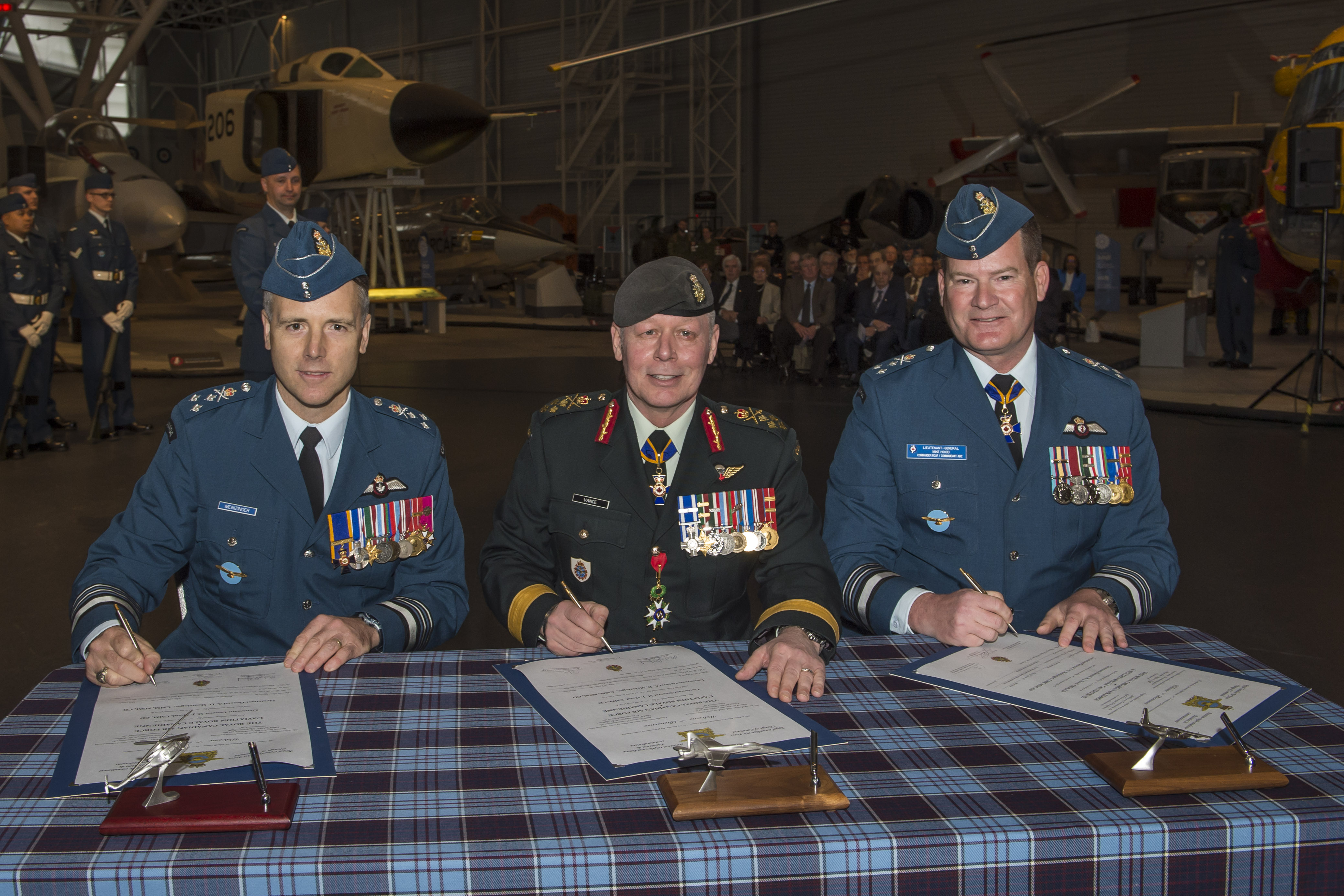 Three men in military uniforms sit behind a tartan-covered table, with documents resting on the table.