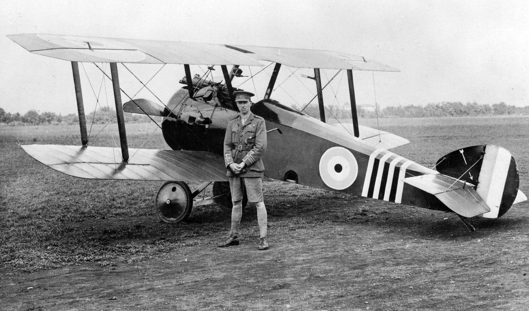 An old black and white photograph of a man in military uniform standing in front of a bi-plane. 