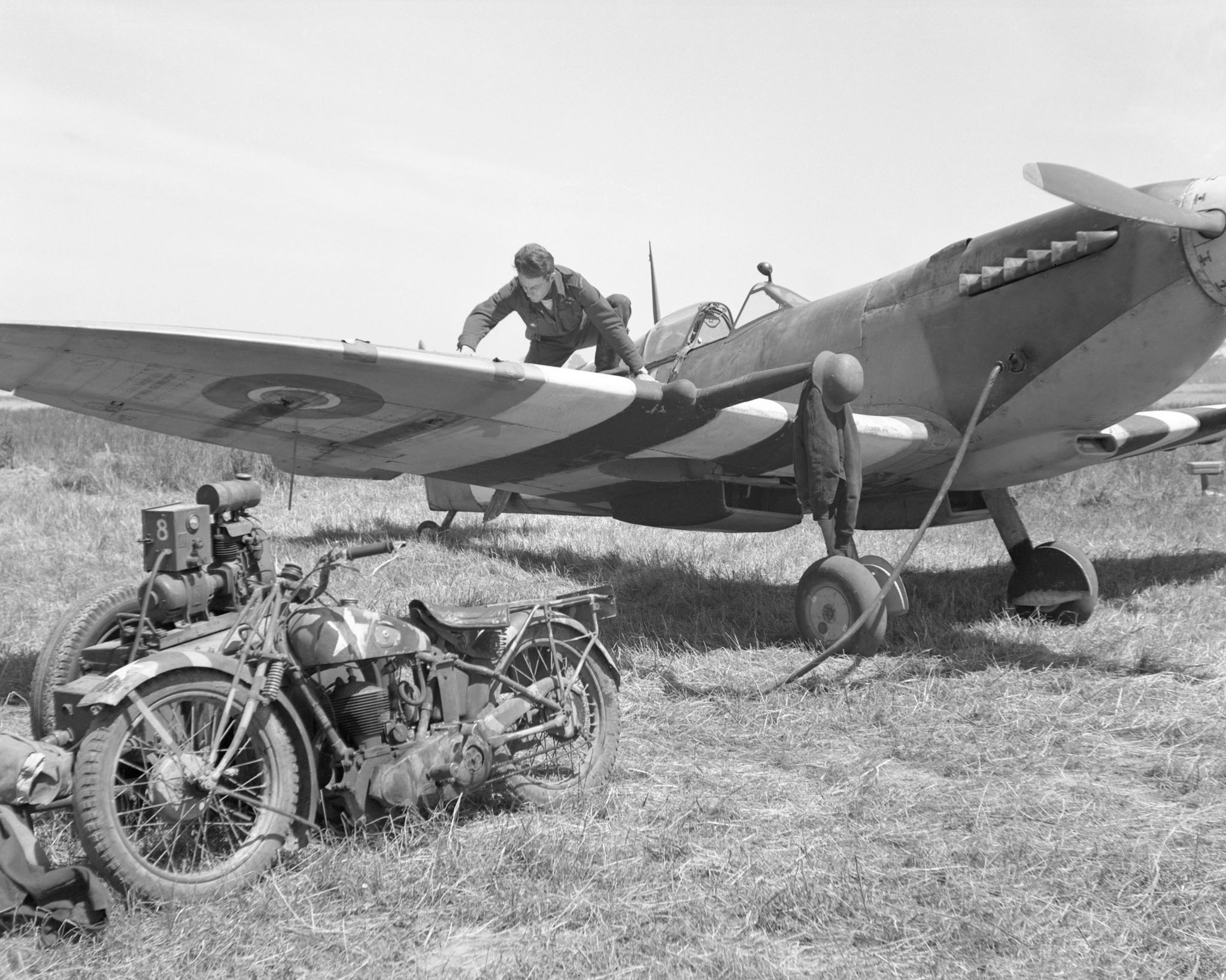 A man crouches on the wing of a propeller-driven fighter aircraft. The wing are painted in stripes. A motorcycle is in the foreground.