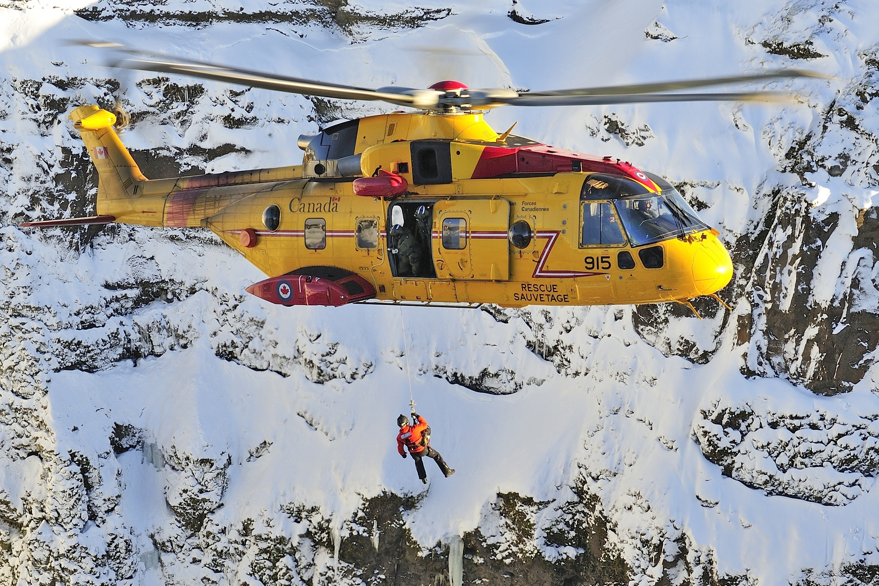 A person hangs suspended on a tether from a large helicopter flying near snow-covered, mountainous terrain.