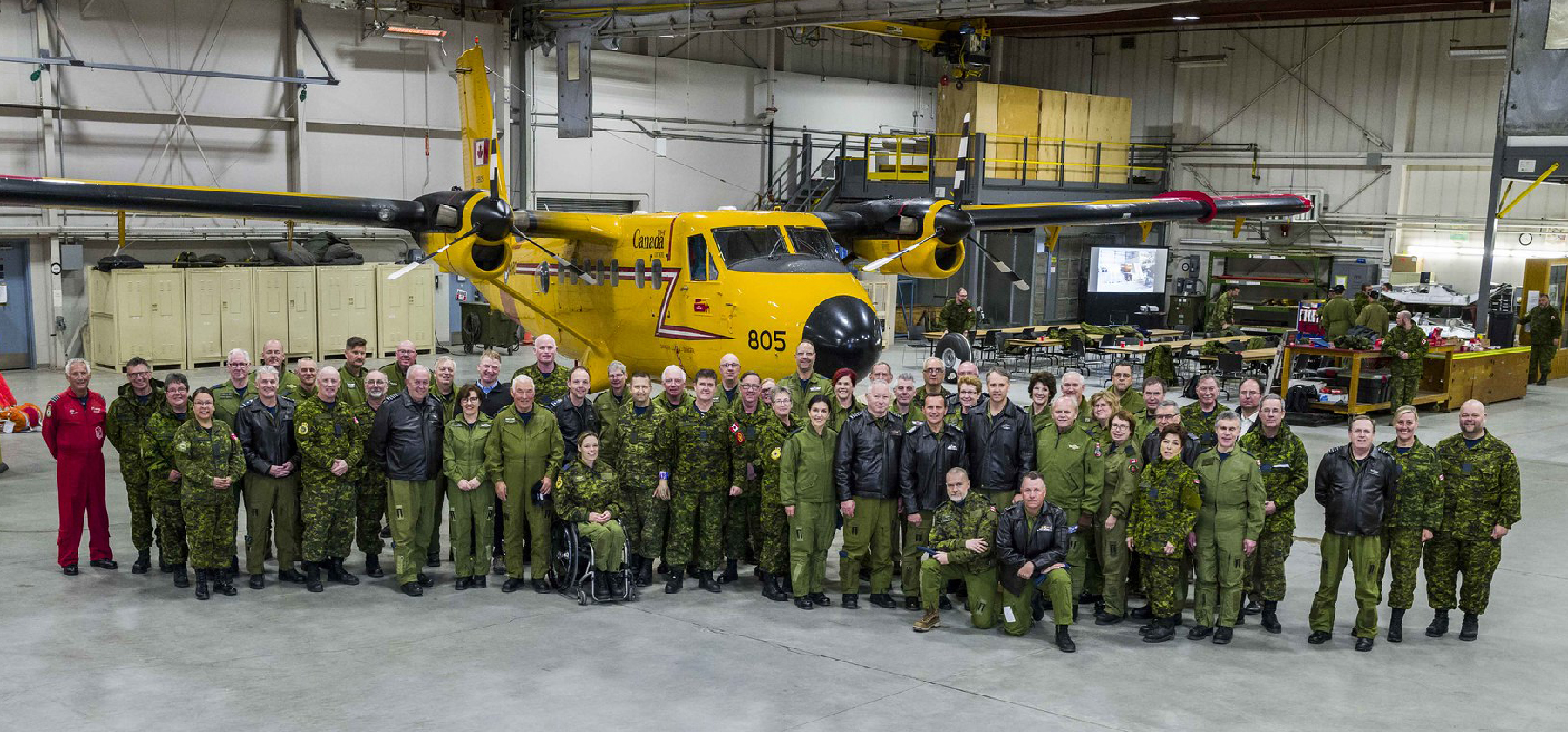 A large group of people, one wearing an orange flightsuit and the rest in olive green, with some in black leather jackets, gather beside a small yellow twin-engine ai\rcraft.