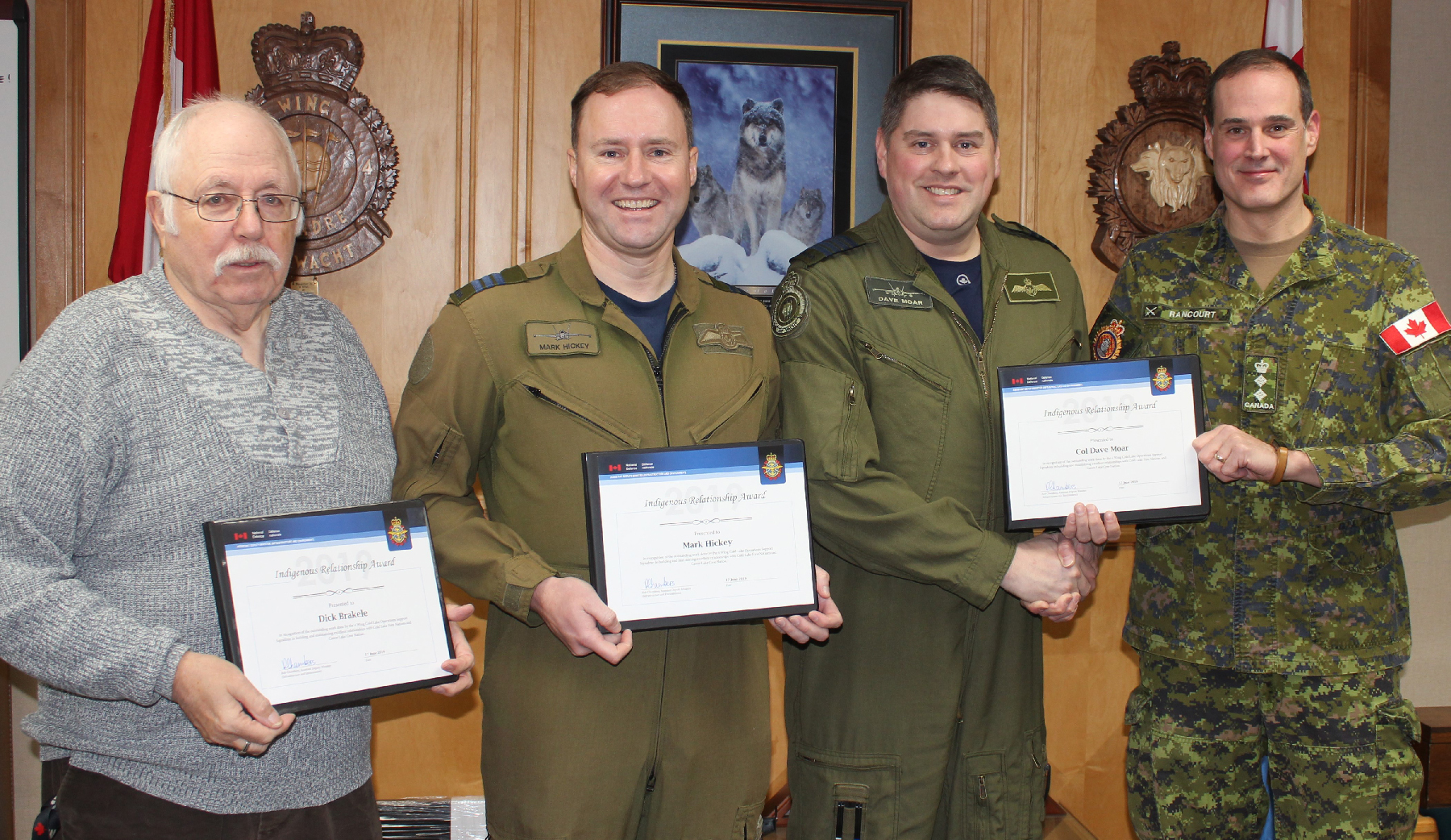 Four men, three in uniform and one in civilian dress, stand in a row holding certificates.