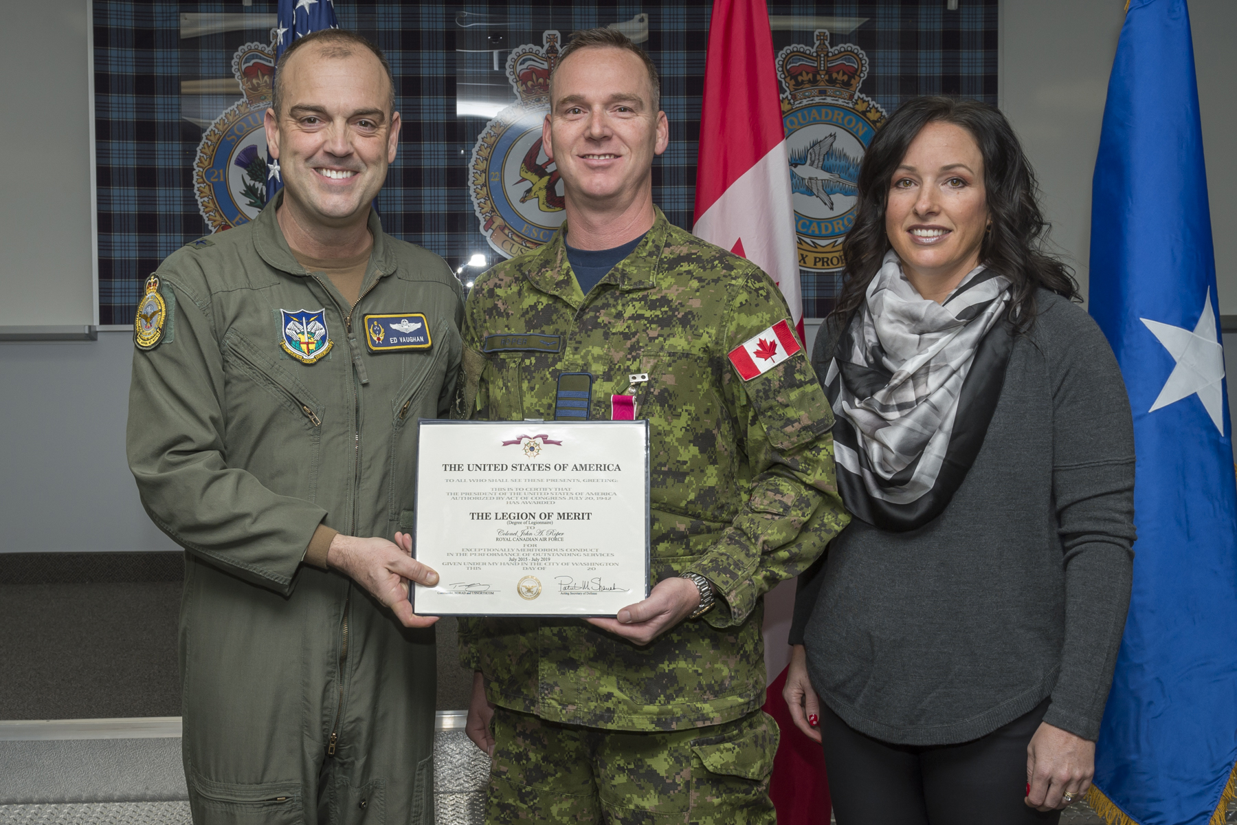 A photo of two men holding a certificate, one wearing a green flight suit and the other a military camouflage uniform, and a woman dressed in civilian clothes.