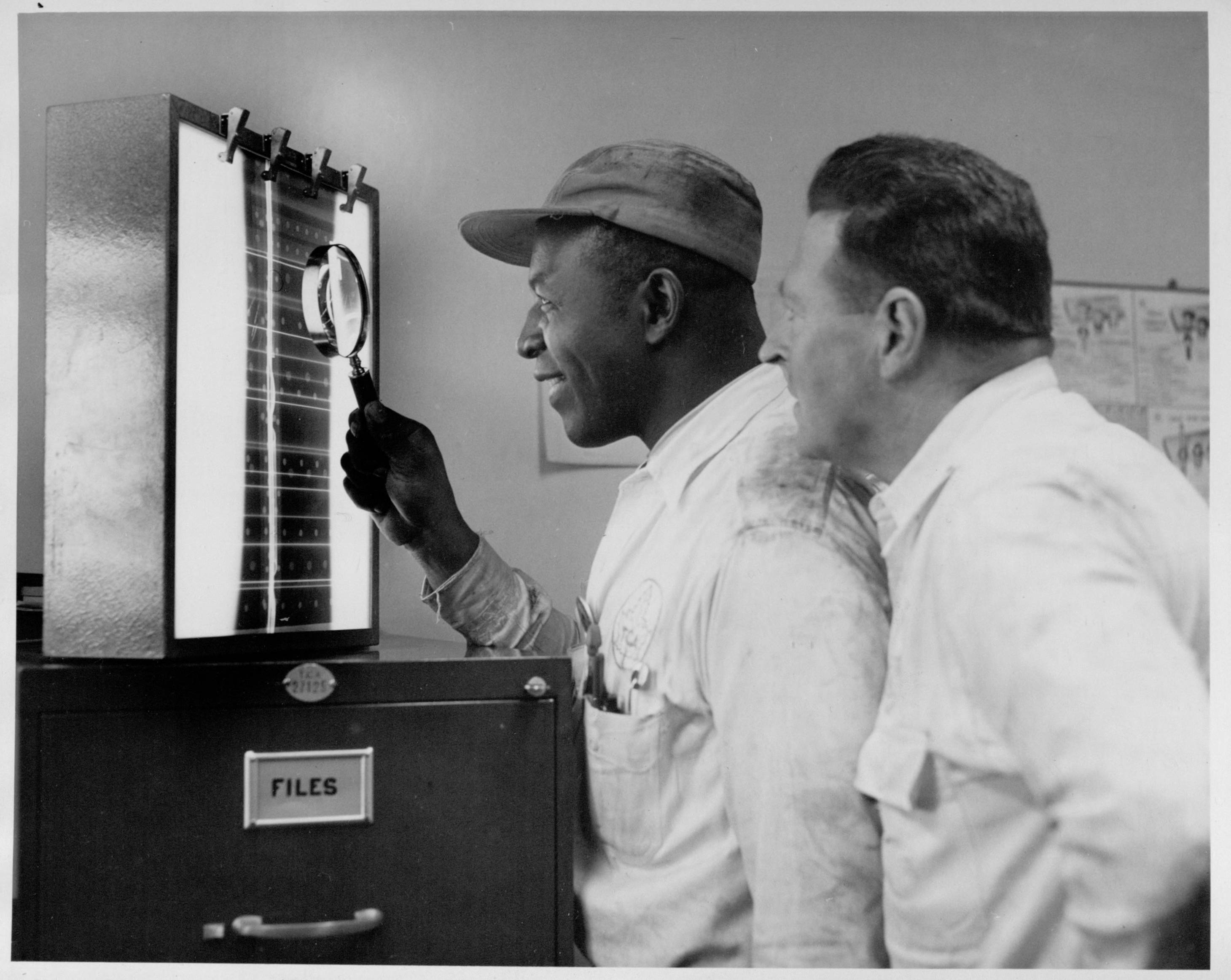 A black and white photo of two men looking at an X-ray film mounted on a viewer, set on top of a file cabinet.