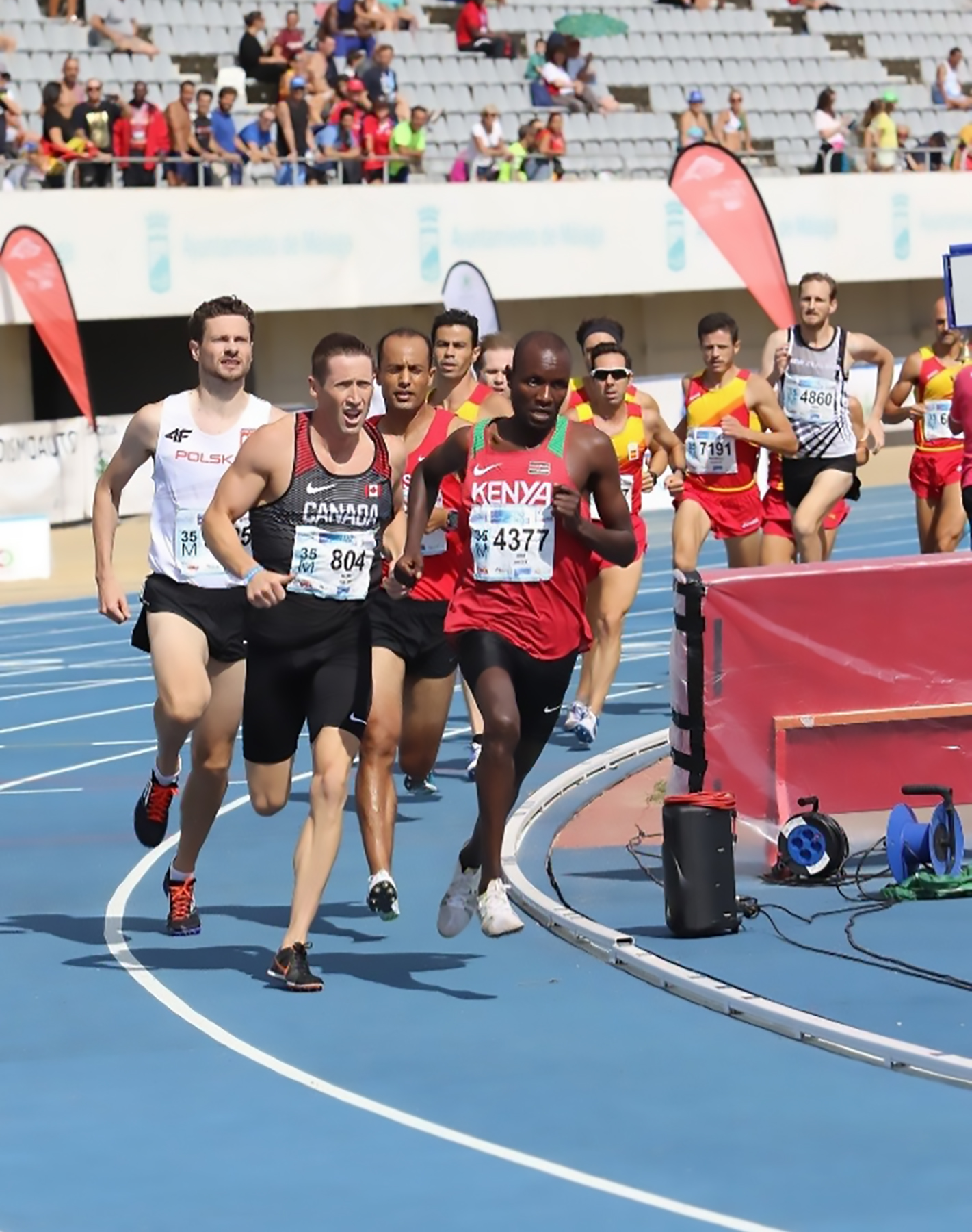 A group of male runners race on an oval track in front of spectators.