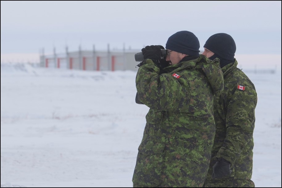 Deux personnes vêtues d’uniformes à camouflage se tenant dans la neige se servent de jumelles.