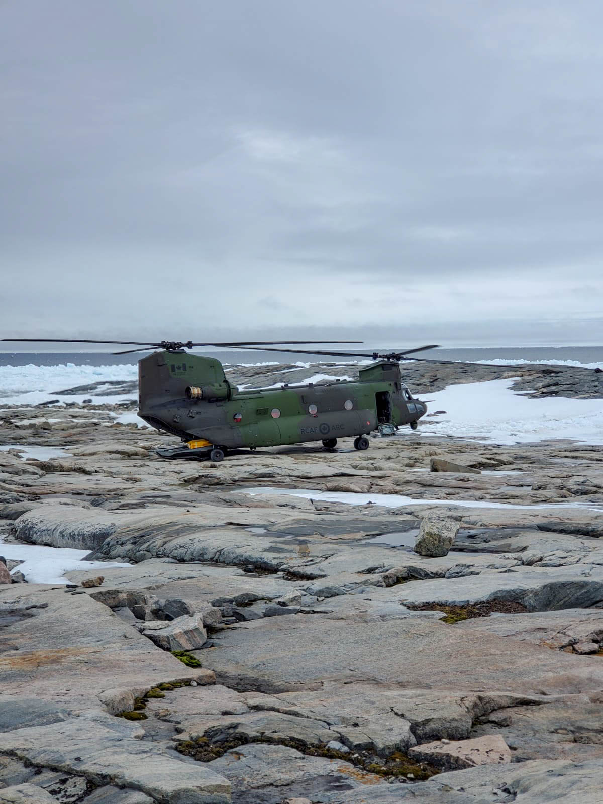 A big green and grey helicopter sits on rocks in an Arctic setting.