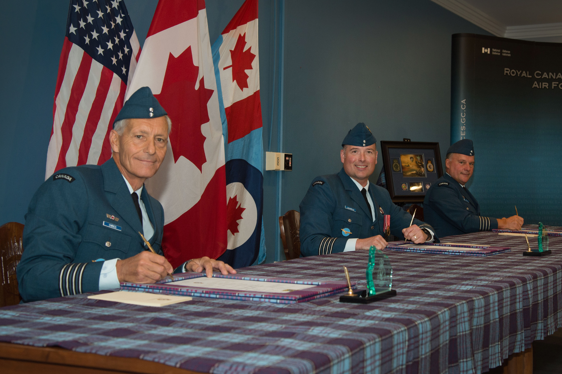 Three men wearing blue military uniforms and holding pens sit at a table covered with a tartan cloth on which there are documents. Behind the men are three flags. 
