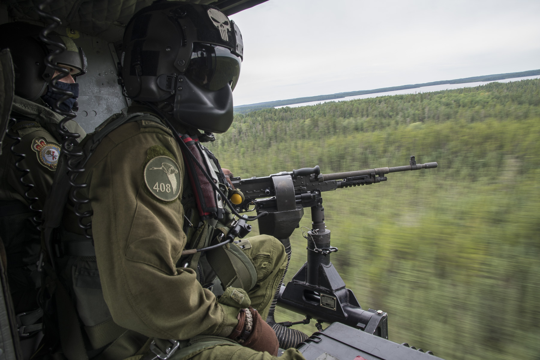Two men wearing green flight suits and black helmets, one of which mans a machine gun, sit at the open door of a helicopter flying over a wooded area.