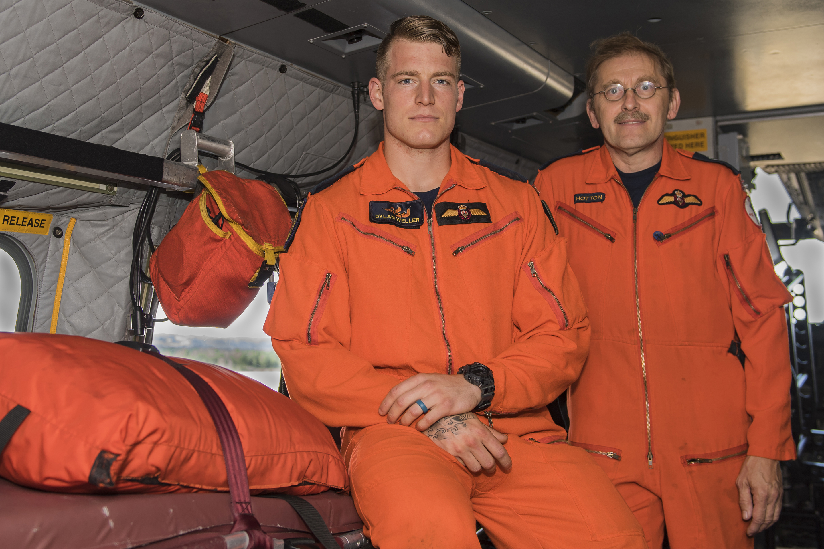 Two men in orange coveralls lean on a bunk in an aircraft.
