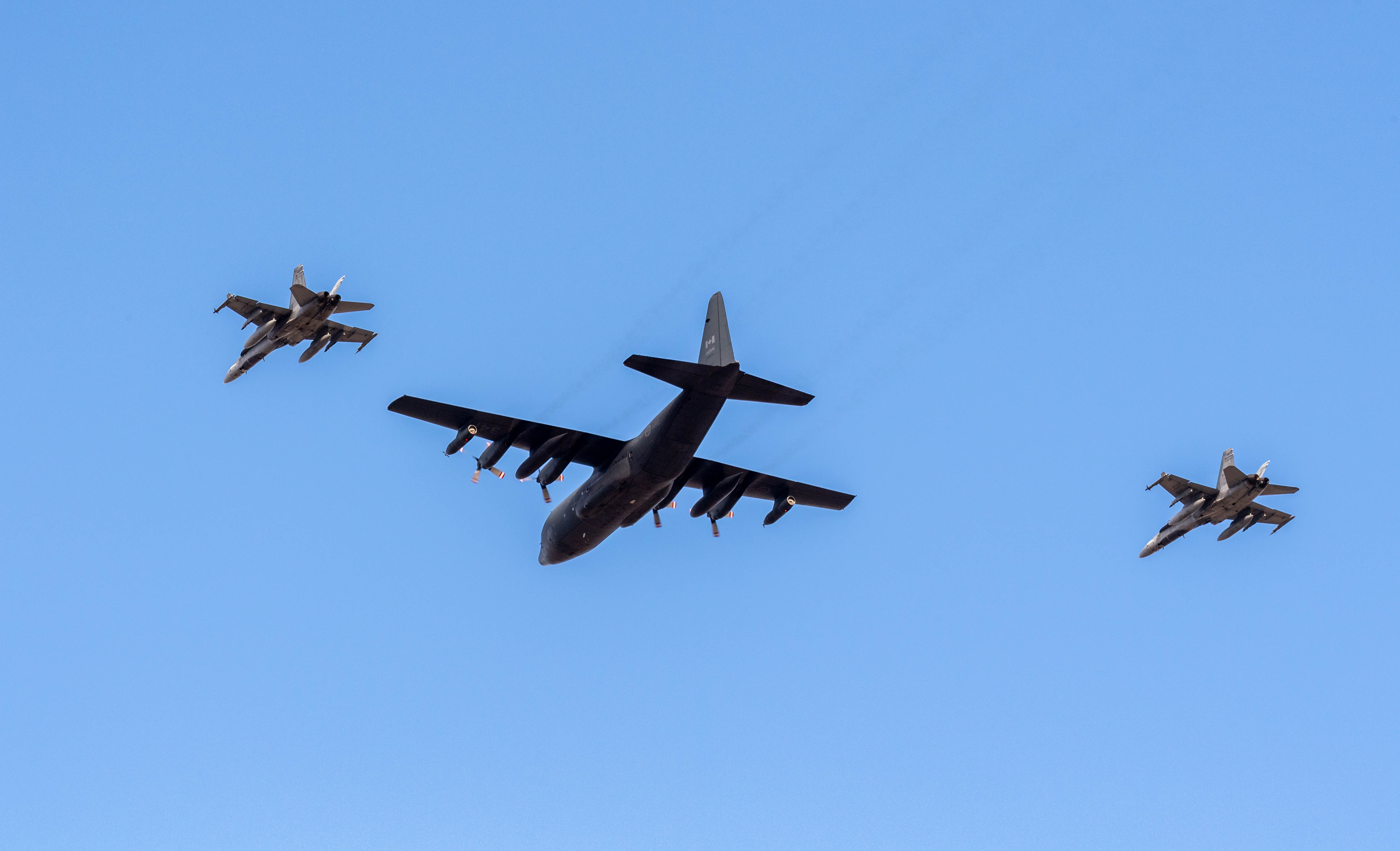 Three aircraft flying in formation in a blue sky. The middle aircraft has propellers and is much bigger that the other two on its sides, which are jet fighters.