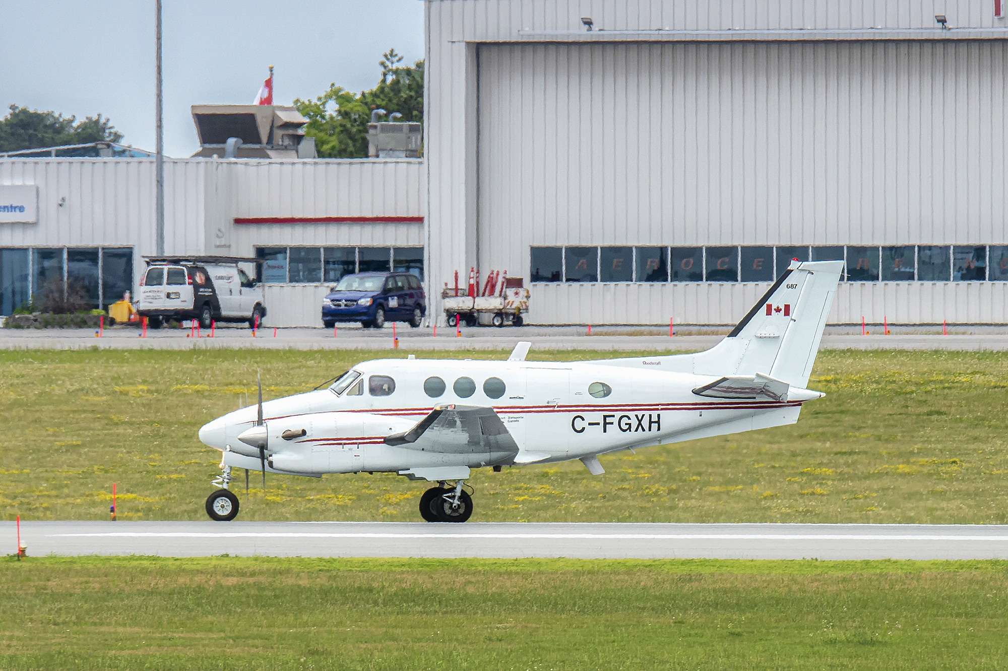 A small white twin propeller aircraft taxis on a runway in front of grey airport buildings.