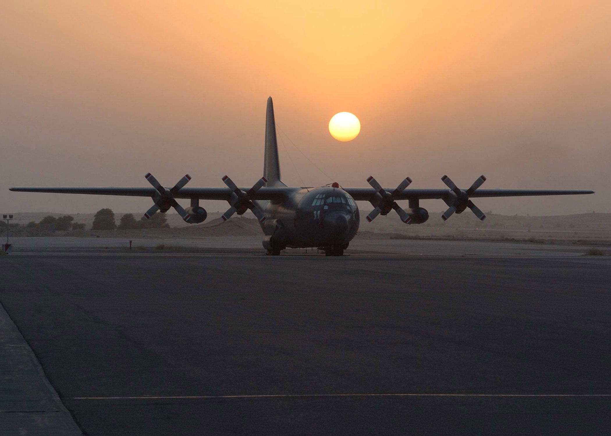 A big four-engine propeller aircraft sits on the tarmac while the sun sets behind it.