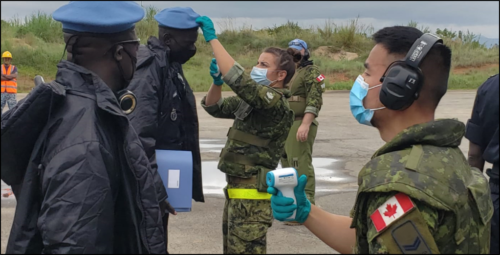 Many people wearing different kinds of military uniforms and masks stand on a runway. Three of those people, who are wearing green camouflage uniforms, are using thermometers to take the temperature of two people wearing dark blue uniforms and blue berets.