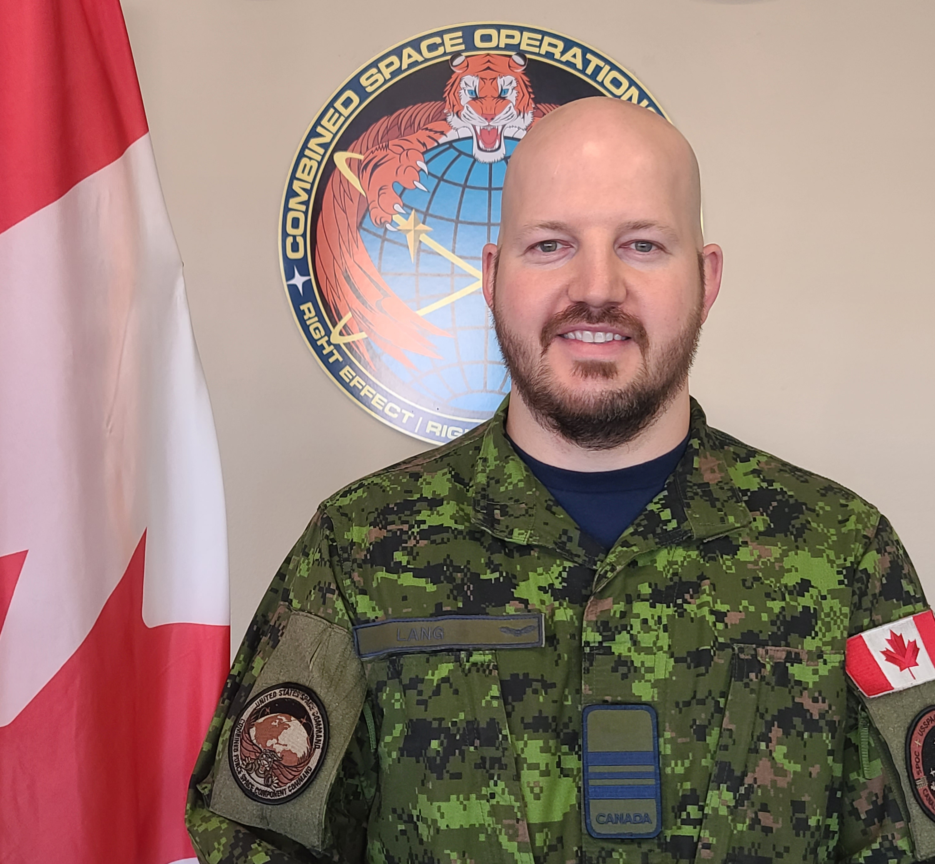 A man wearing a military camouflage uniform stands in front of a Canadian flag and military crests.