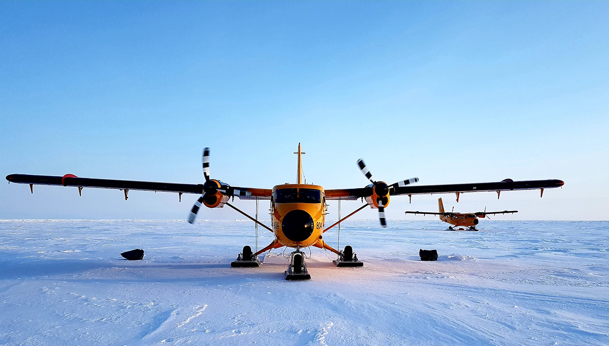 Deux avions orange munis de deux moteurs à hélices et de skis se trouvent sur la neige dans l’Arctique.