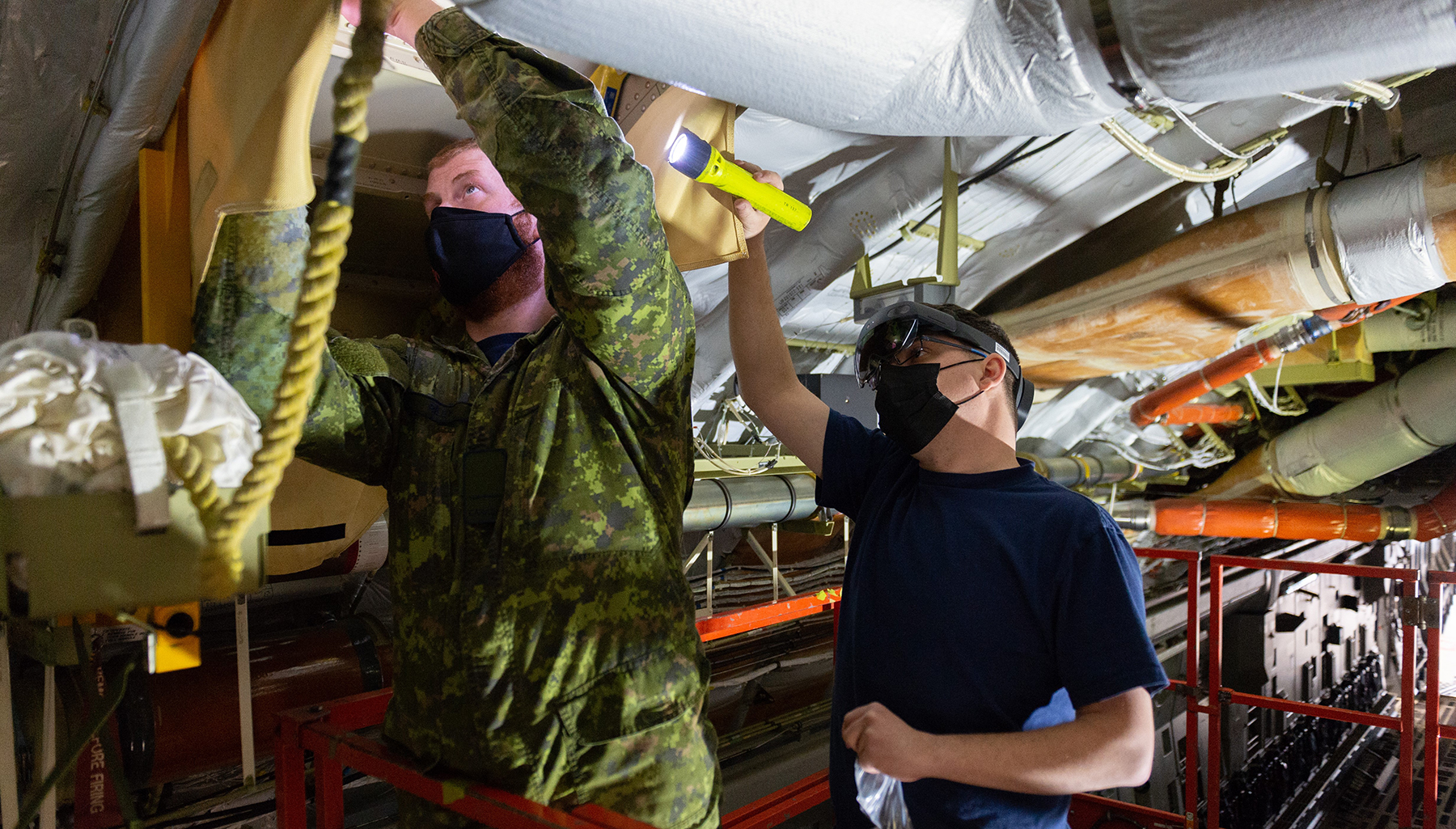 Two men with masks, one wearing an olive-green camouflage uniform and the other a blue shirt, work inside an aircraft. 