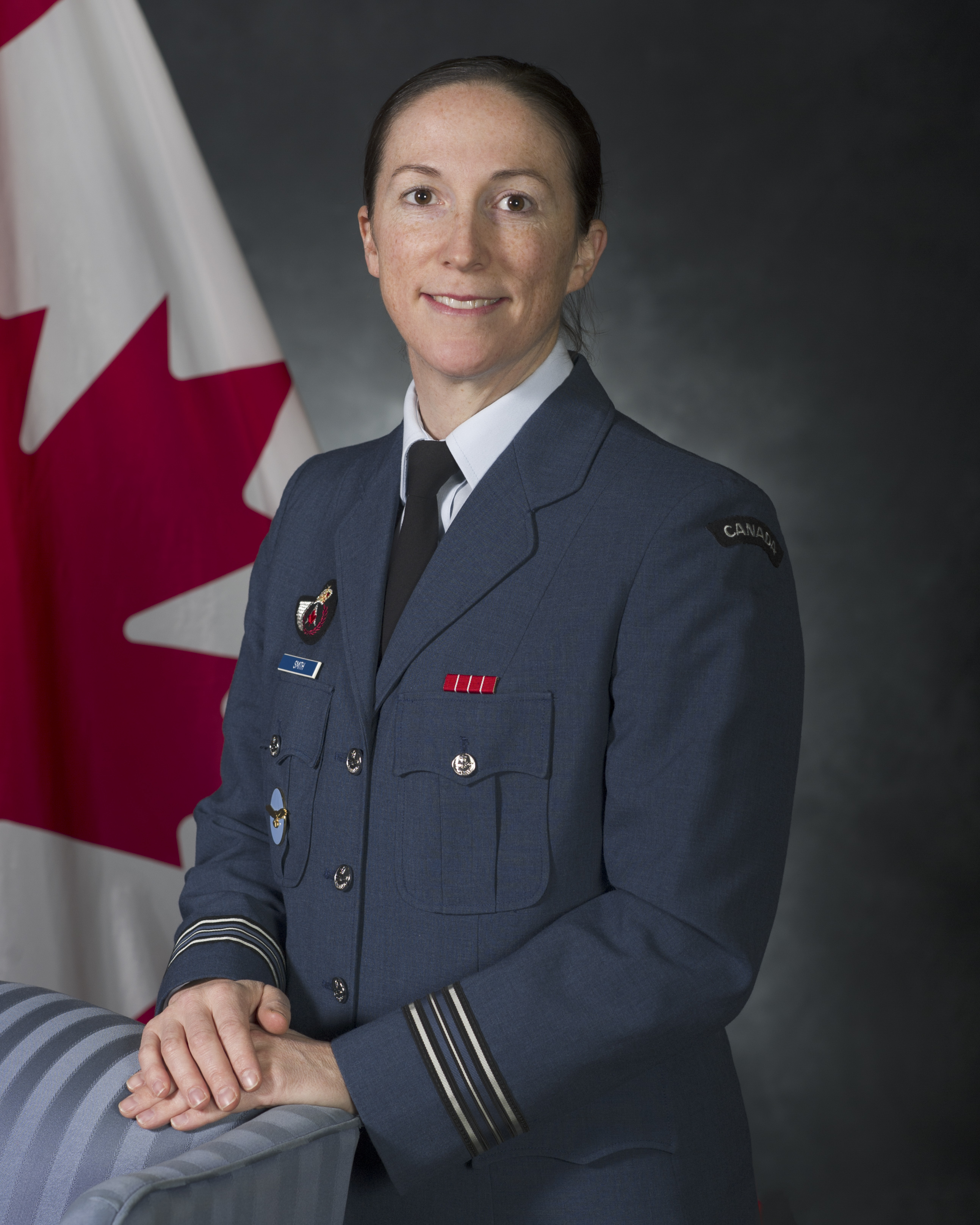A smiling woman wearing a blue military uniform stands in front of a Canadian flag, her hands resting on a blue chair.